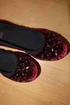 A pair of burgundy velvet ballet flats decorated with black and brown sequins and beads, shown on a light wood floor.