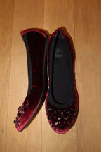 A pair of burgundy velvet ballet flats with black beaded embellishments on the toes, displayed on a light wood floor.