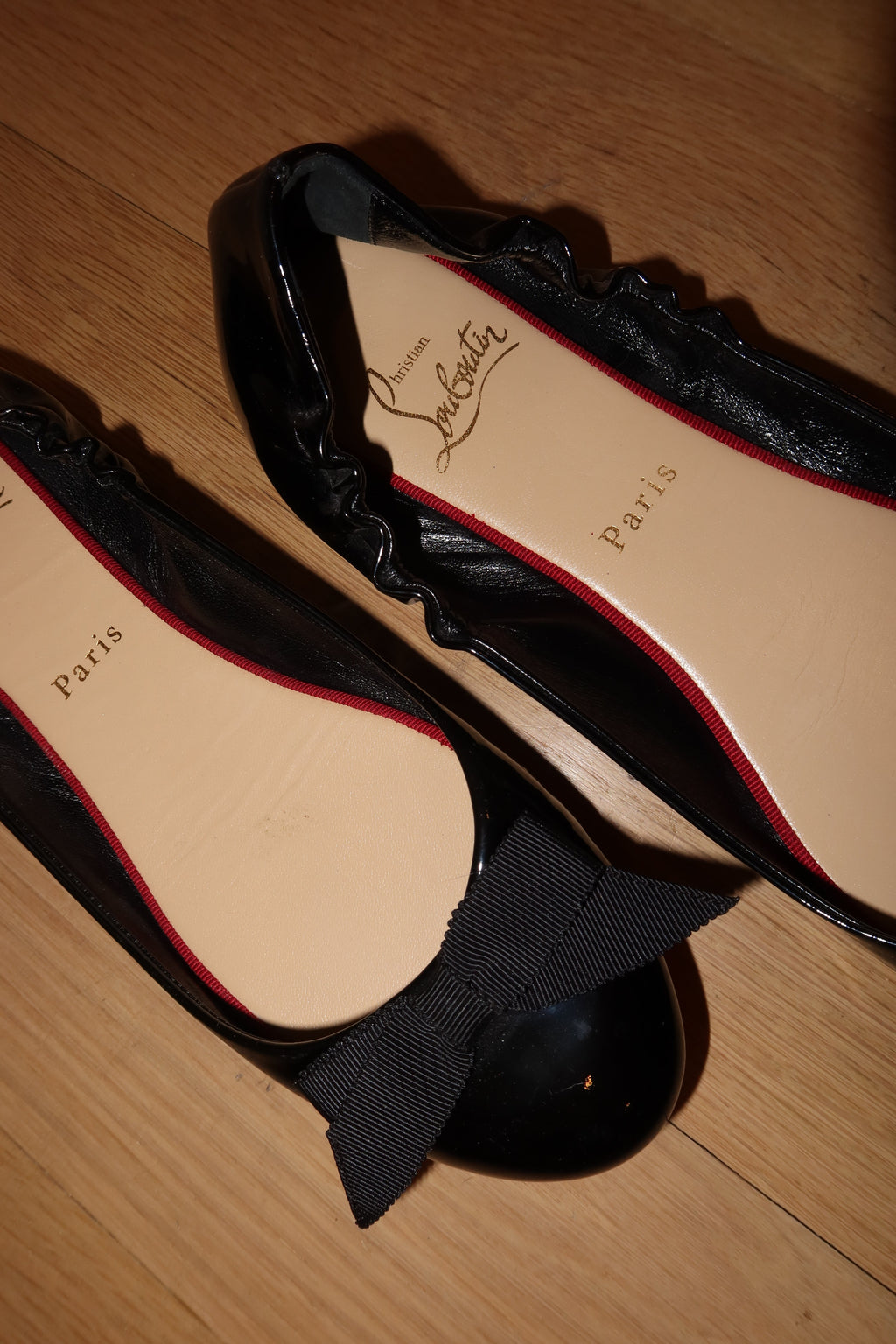 A pair of black patent leather Christian Louboutin ballet flats with a bow on one shoe and ruffle trim on the other, displayed on a wooden floor. The beige insoles feature the 'Christian Louboutin Paris' logo.