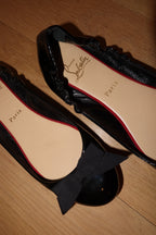 A pair of black patent leather Christian Louboutin ballet flats with a bow on one shoe and ruffle trim on the other, displayed on a wooden floor. The beige insoles feature the 'Christian Louboutin Paris' logo.