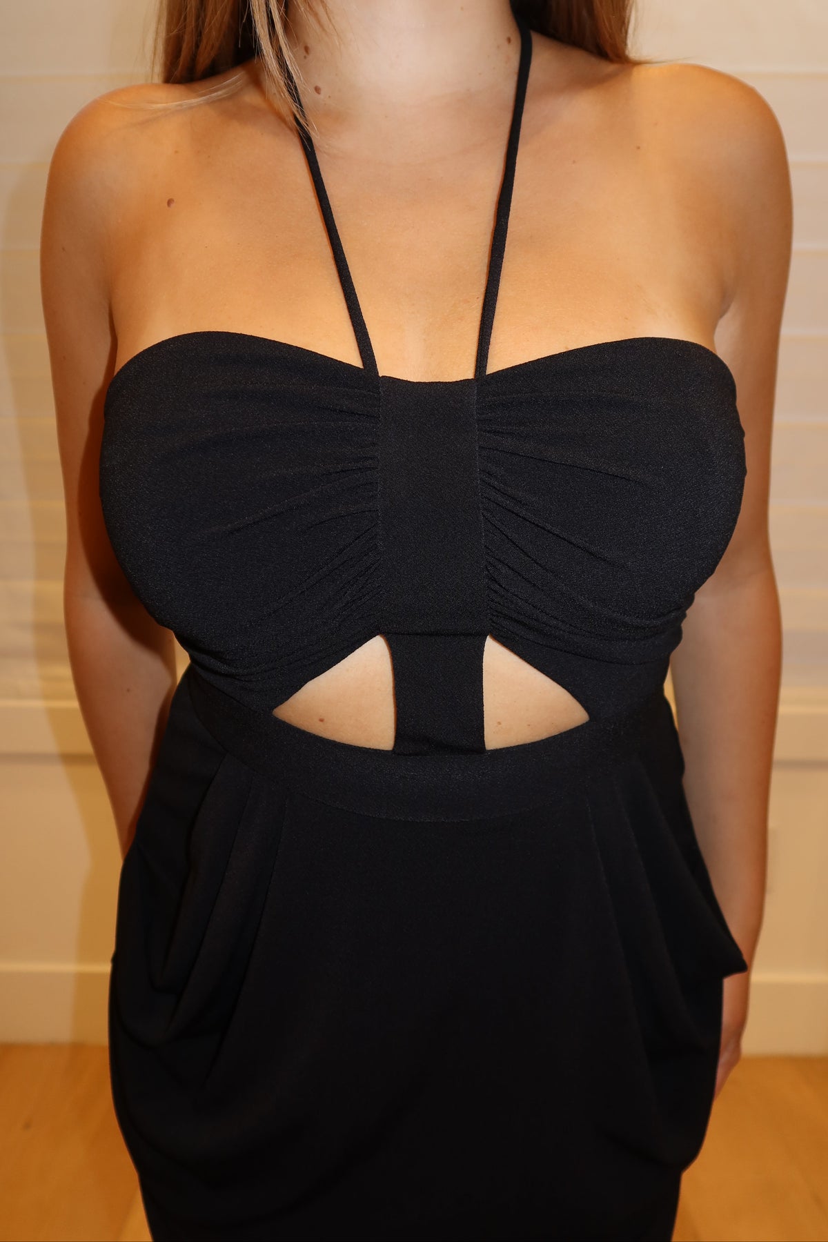 A woman wearing a black strapless halter dress with a sweetheart neckline, gathered bust, and a cutout at the waist, photographed indoors against a light beige paneled wall.