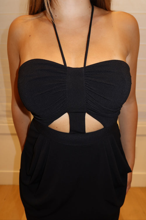 A woman wearing a black strapless halter dress with a sweetheart neckline, gathered bust, and a cutout at the waist, photographed indoors against a light beige paneled wall.