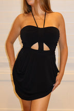 A woman modeling a black strapless mini dress with a sweetheart neckline and a central cutout, standing against a white paneled wall on a light wood floor.