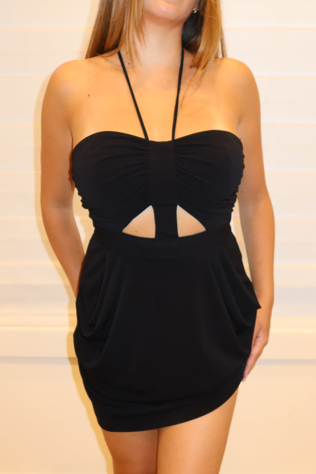 A woman modeling a black strapless mini dress with a sweetheart neckline and a central cutout, standing against a white paneled wall on a light wood floor.