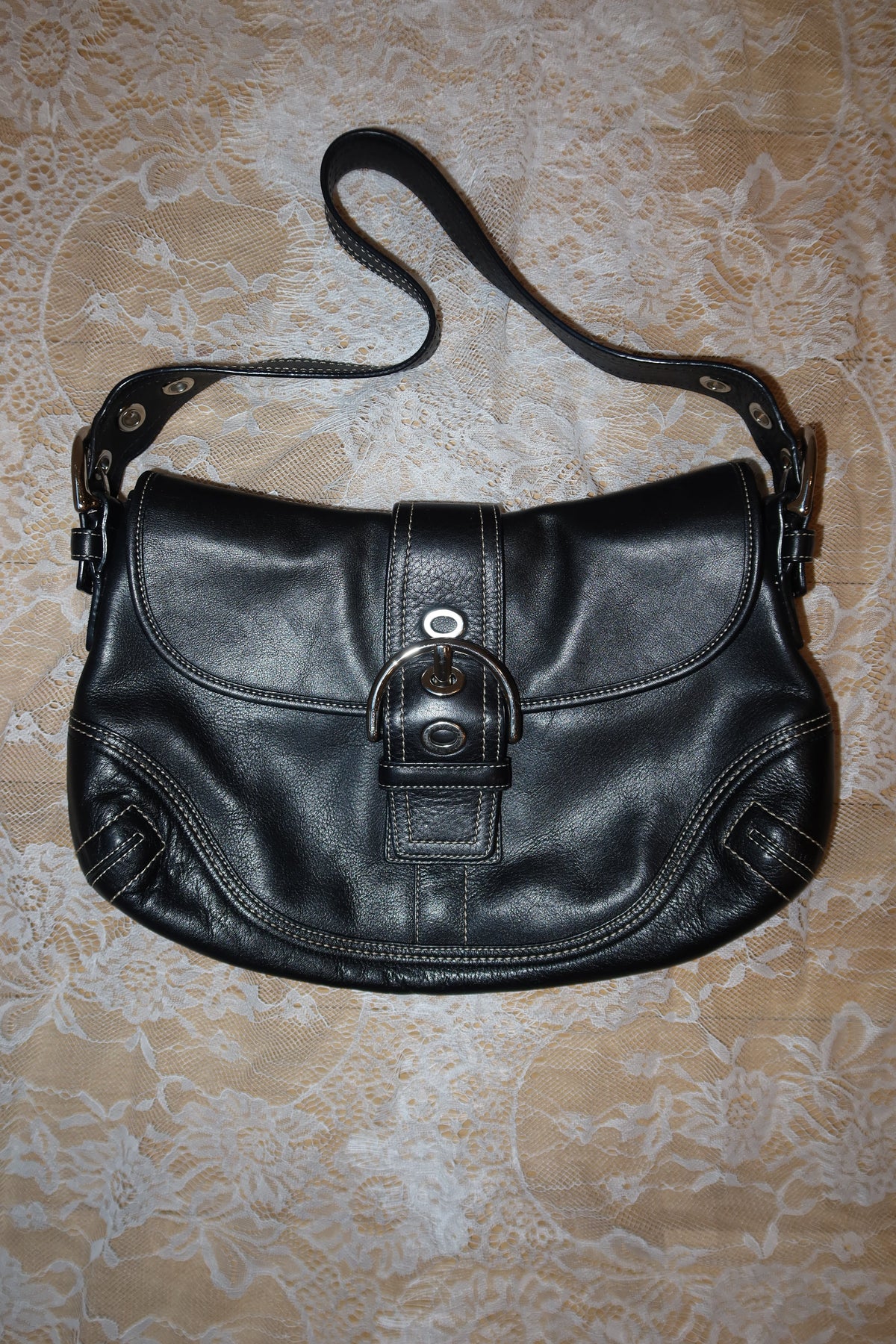 A black leather shoulder bag with a curved flap, a central buckle strap, and a long shoulder strap, laid flat on a beige patterned fabric background.