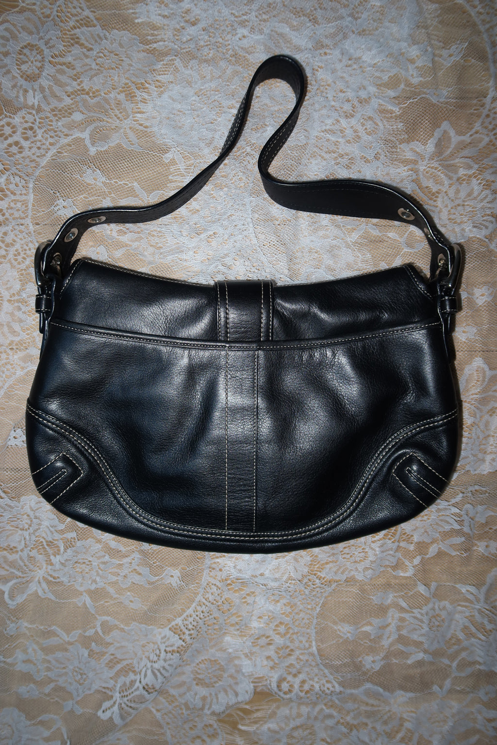 A black leather shoulder bag with a curved crescent shape, laid flat on a beige and white patterned lace fabric background.