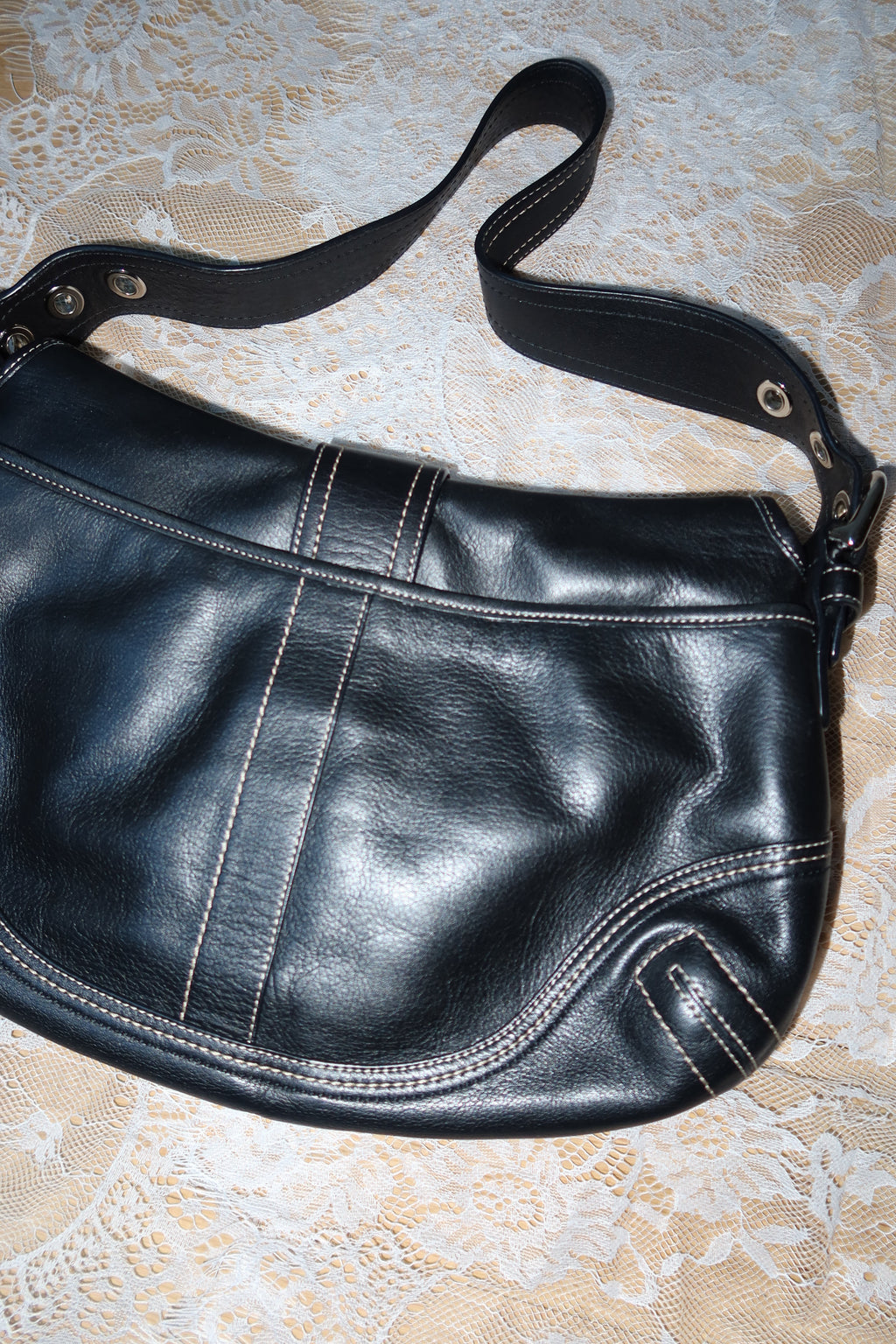 Black leather shoulder bag with white contrast stitching, laid flat on a beige lace fabric background.