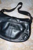 Black leather shoulder bag with white contrast stitching, laid flat on a beige lace fabric background.