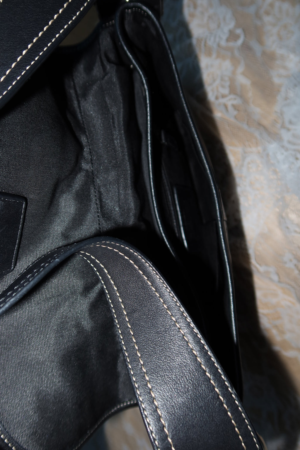 Close-up of a black leather or faux-leather bag interior with visible white contrast stitching and a dark fabric lining, resting on a patterned beige surface.