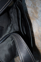 Close-up of a black leather or faux-leather bag interior with visible white contrast stitching and a dark fabric lining, resting on a patterned beige surface.