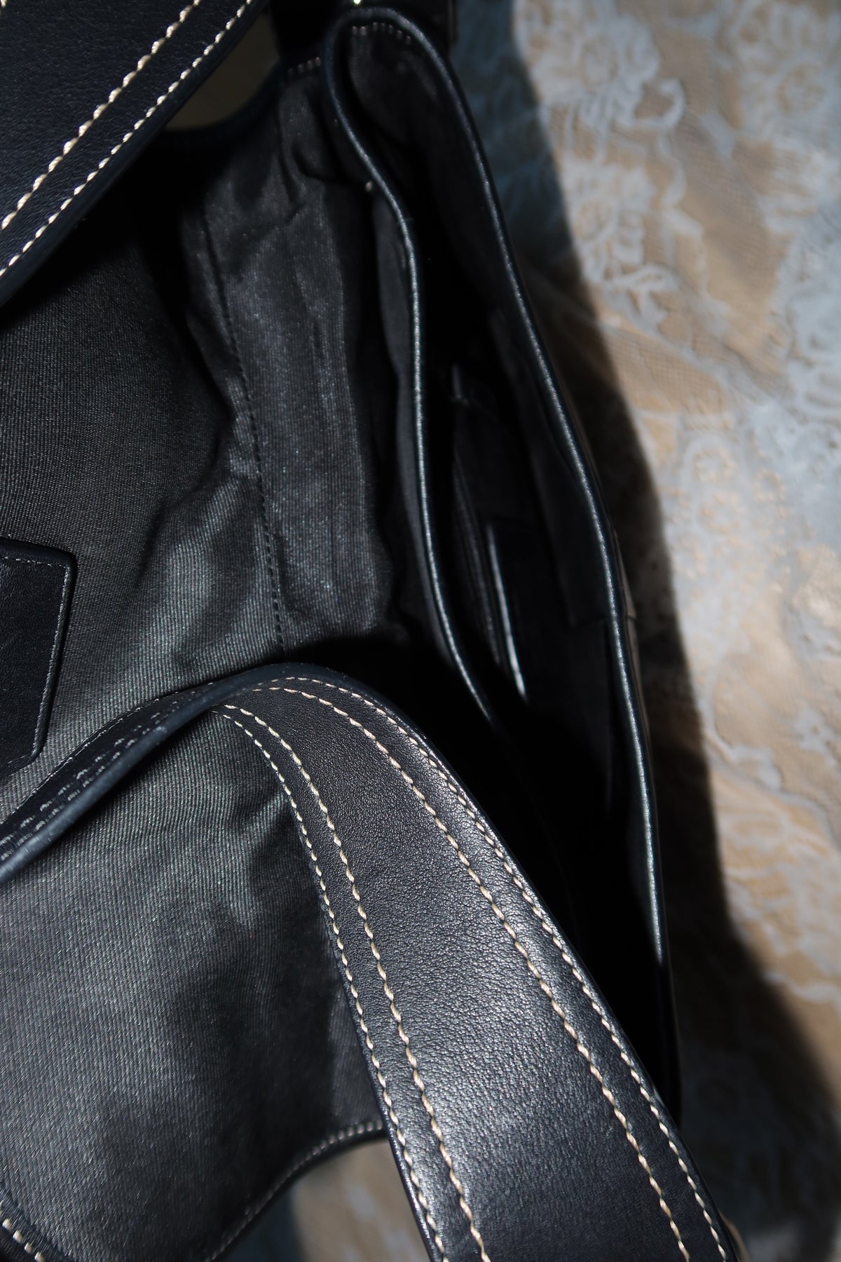 Close-up of a black leather or faux-leather bag interior with visible white contrast stitching and a dark fabric lining, resting on a patterned beige surface.
