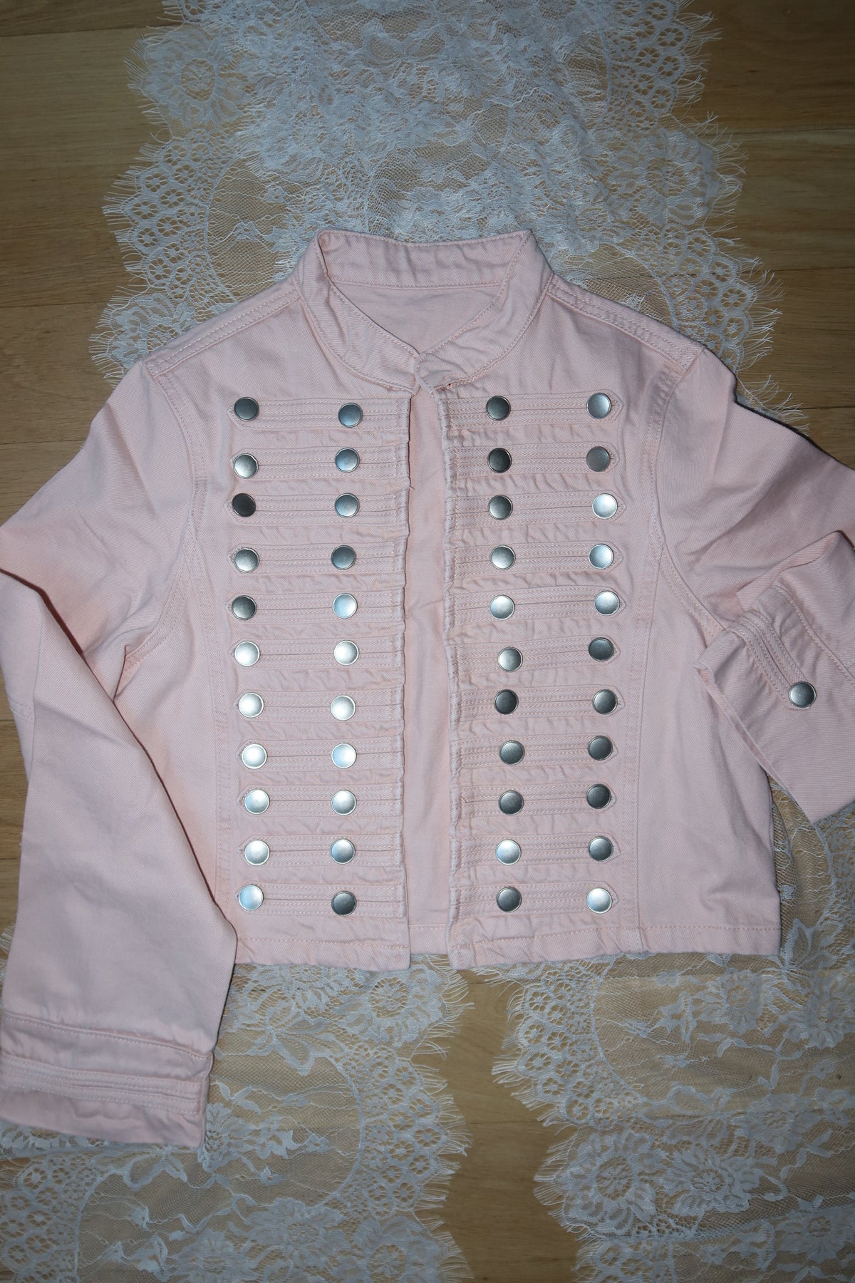 A light pink long-sleeve top with rows of silver metallic studs arranged in horizontal lines, laid flat on a white lace sheet over a wooden surface.