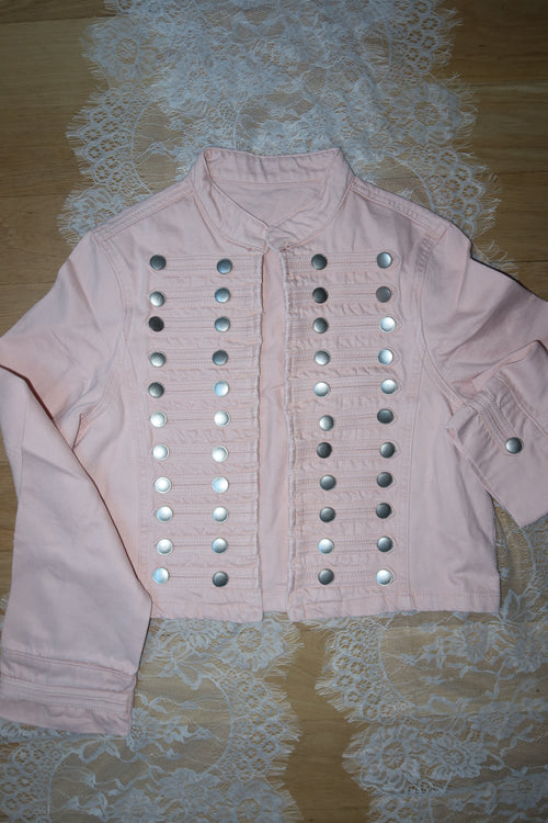 A light pink long-sleeve top with rows of silver metallic studs arranged in horizontal lines, laid flat on a white lace sheet over a wooden surface.