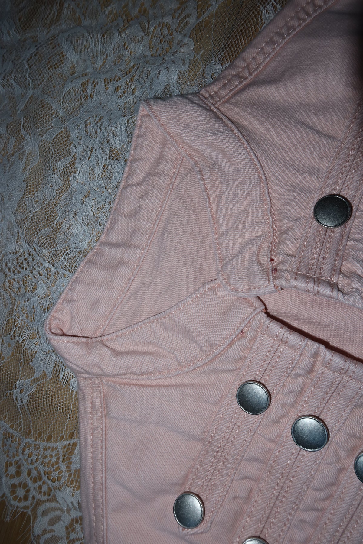 A close-up of a light pink denim jacket featuring silver snap buttons and a pointed collar, laid flat on a beige lace fabric background.